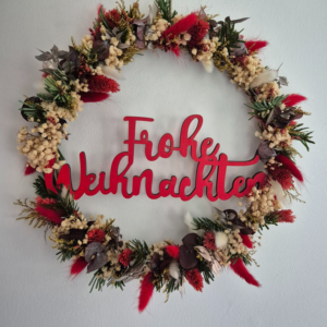 Blumenring mit einem Schriftzug aus Spanplatte in rot mit der Aufschrift Frohe Weihnachten. Der Ring ist mir Trockenblumen umringt.