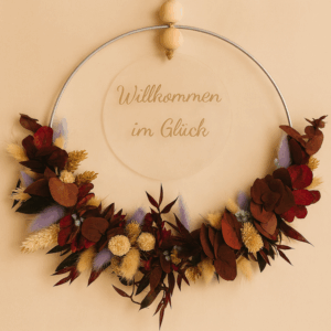 Blumenkranz, der untere Teil ist mit Trockenblumen bedeckt. In der Mitte ist mit einem Juteband eine Acrylplatte mit der Aufschrift "Willkommen im Glück".