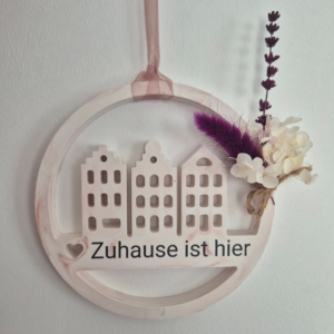 Loop mit Häusern in der Mitte und der Aufschrift "Zuhause ist hier", abgerundet von einem kleinen Strauß Trockenblumen